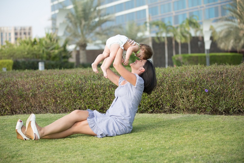 Mother's Day Gifts Mom Will Love a Mom Sitting on a Lawn at a Park Holding her Baby Up Above Her Face
