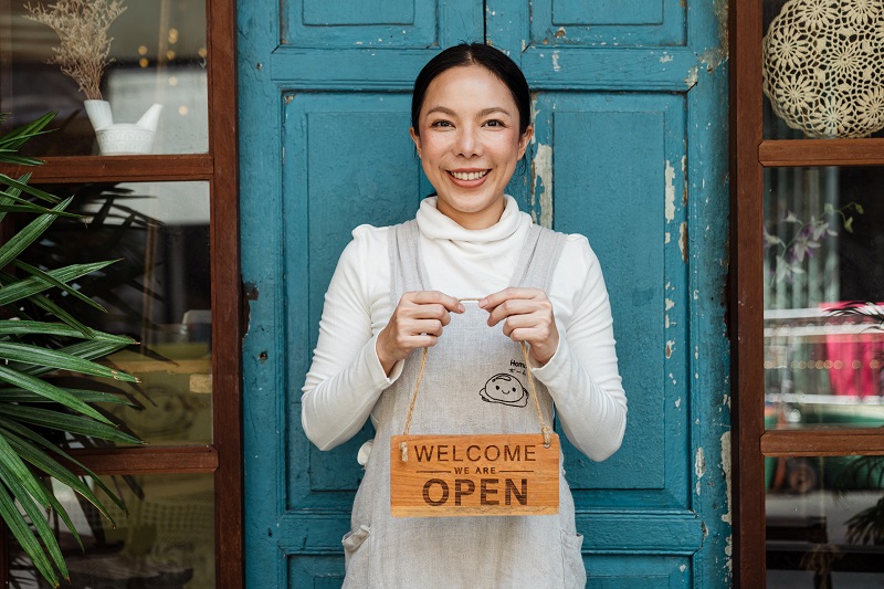 How to Lower Your Small Business Electricity Bill Woman Standing in Front of a Door Holding Up an Open Sign
